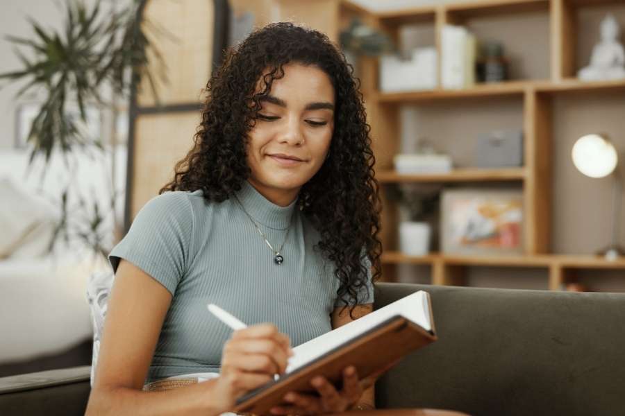 Young woman with curly hair reading book that contains creative writing prompts in modern living space