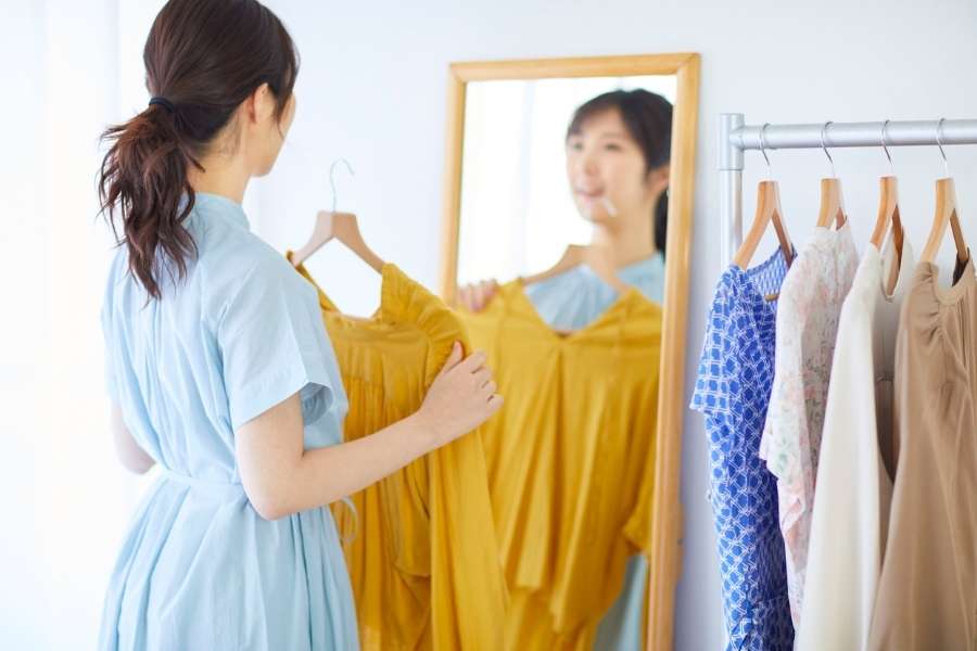 Woman in blue dress selecting yellow garment while looking in mirror with clothing rack